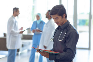 Side view of Caucasian male doctor looking at medical file in hospital. In the background diverse colleagues are discussing in the hallways.