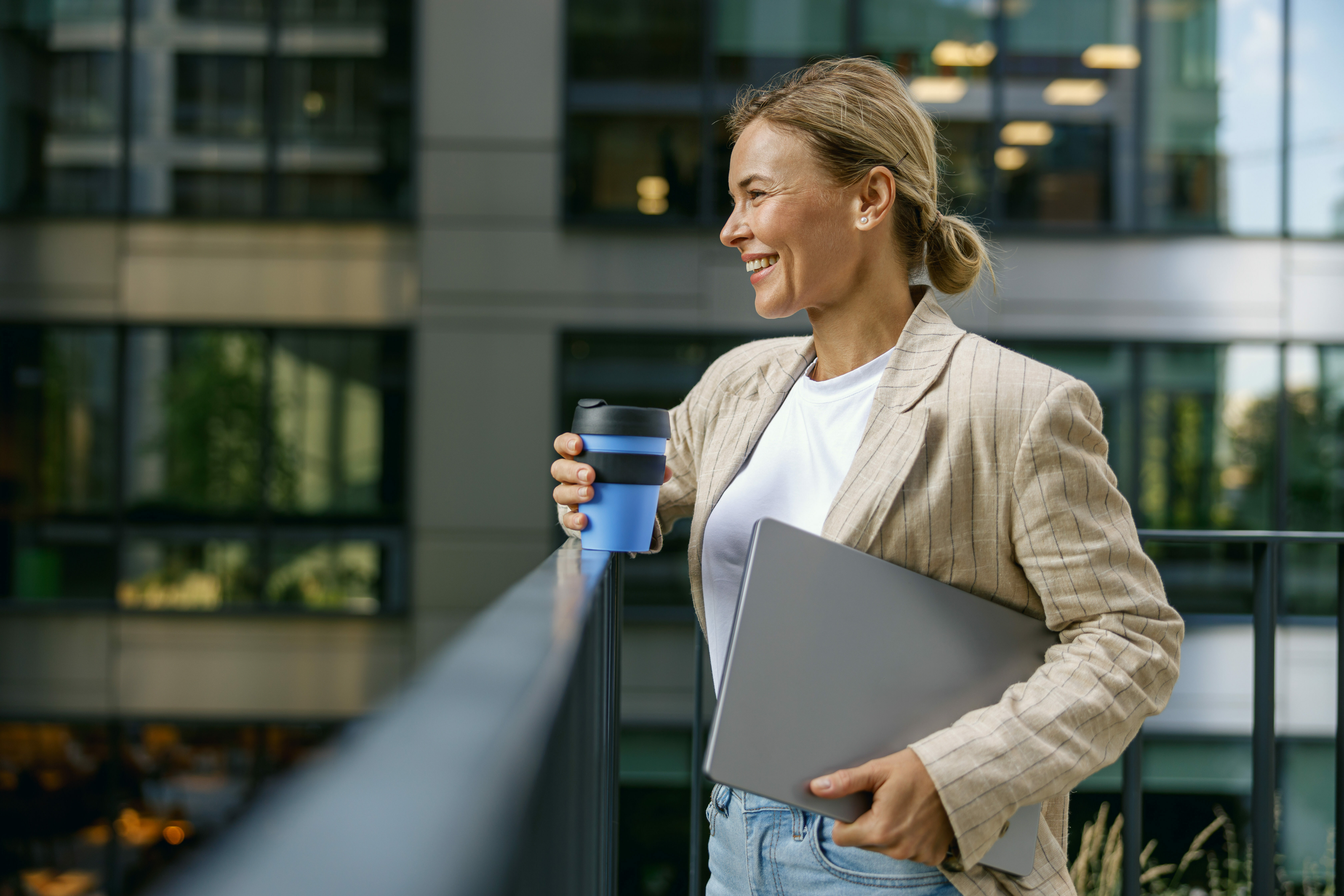 Woman freelancer standing with laptop and coffee cup on modern building background during break time