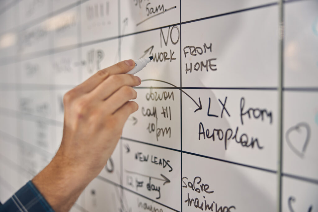 Close up of male hand filling in planner board in office - Image depicting project workflow with a pen, highlighting care coordination, healthcare communication, integrated healthcare, and patient management software.