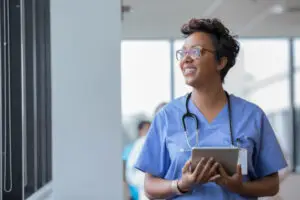 Female nurse holding a digital tablet, smiling while looking out the window, highlighting patient management software, care coordination, healthcare communication, and integrated healthcare.